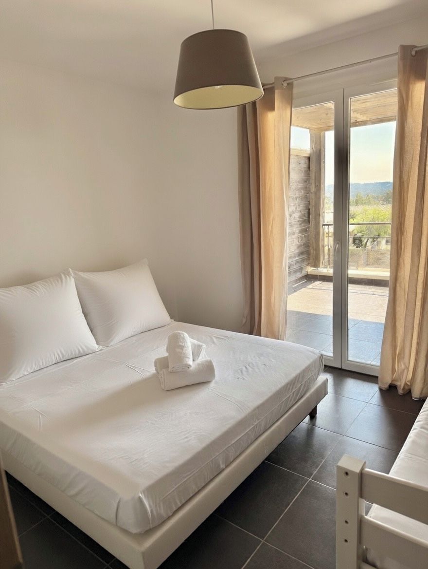 A hotel room with a bed, a hanging light fixture, a view through a glass door, and a balcony.