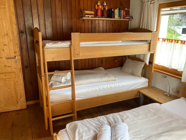 Quarto com beliche, paredes de madeira, uma estante com livros e miniaturas, e janela com cortinas.