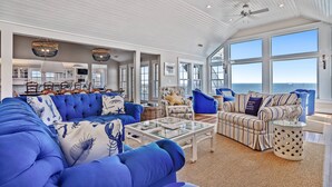 A spacious living area with a large blue sofa, a striped sofa, and a glass coffee table. There are multiple windows providing a view of the ocean. - Frankly My Dear - Duck, Oceanfront, Elevator, Wheelchair Friendly, Pool, Hot Tub, Flex Stays (Duck)