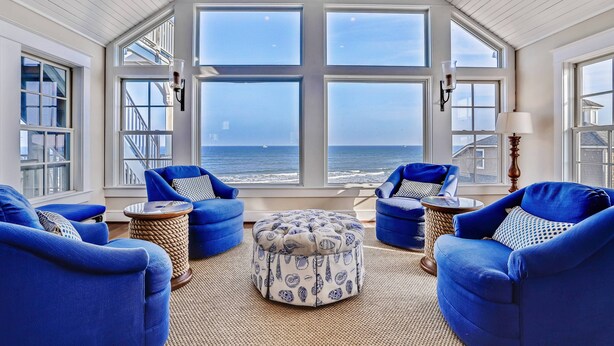 A room with large windows, blue upholstered chairs, a round ottoman, and a lamp. - Frankly My Dear - Duck, Oceanfront, Elevator, Wheelchair Friendly, Pool, Hot Tub, Flex Stays (Duck)