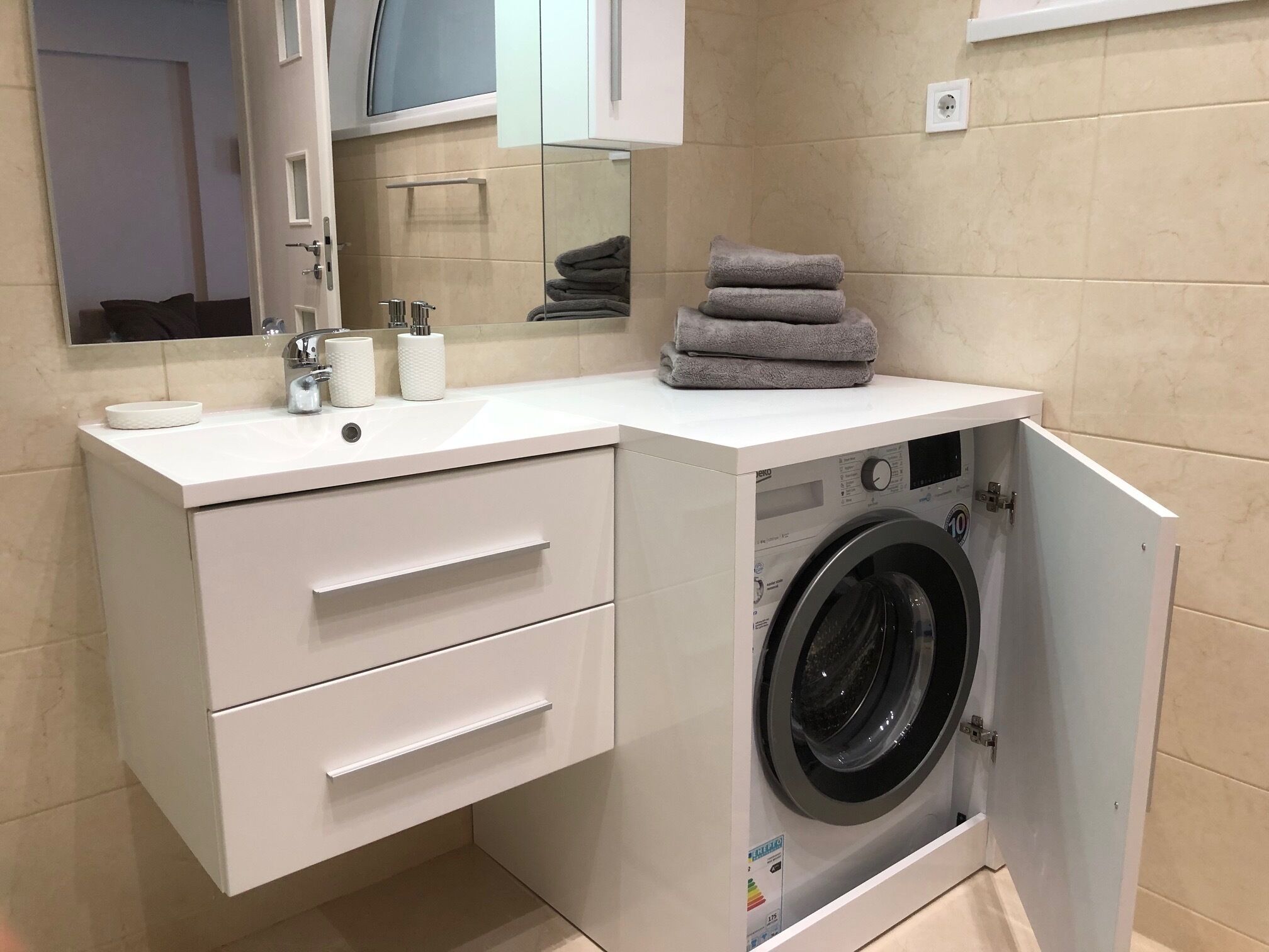 A compact bathroom with a built-in washing machine, a sink with toiletries, and a mirror.