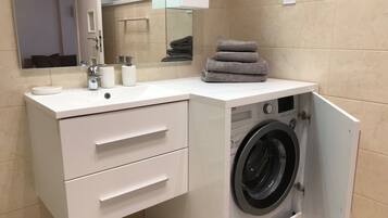 A compact bathroom with a built-in washing machine, a sink with toiletries, and a mirror.