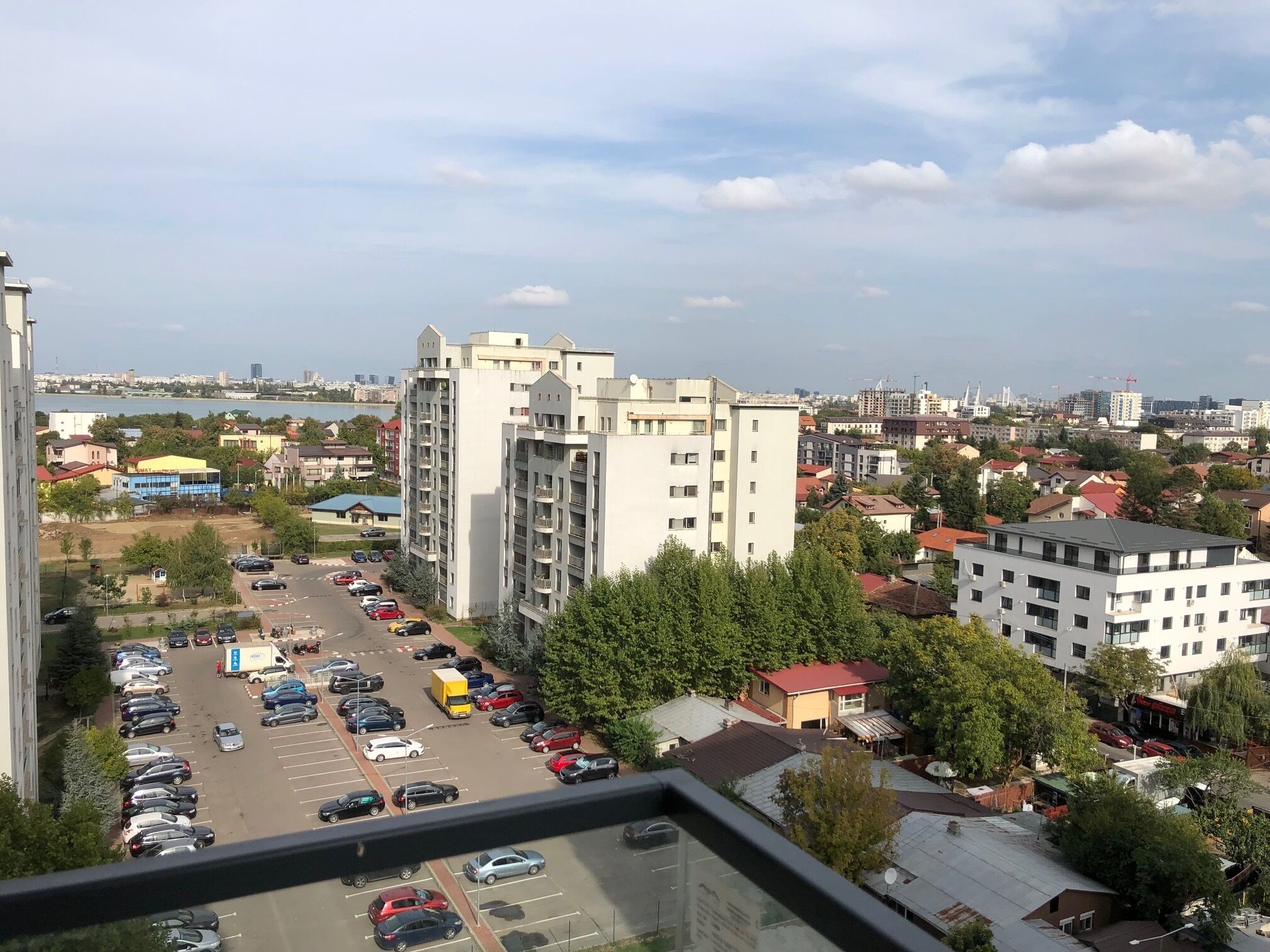 A view of a parking lot with several cars and a yellow truck, surrounded by buildings and trees.