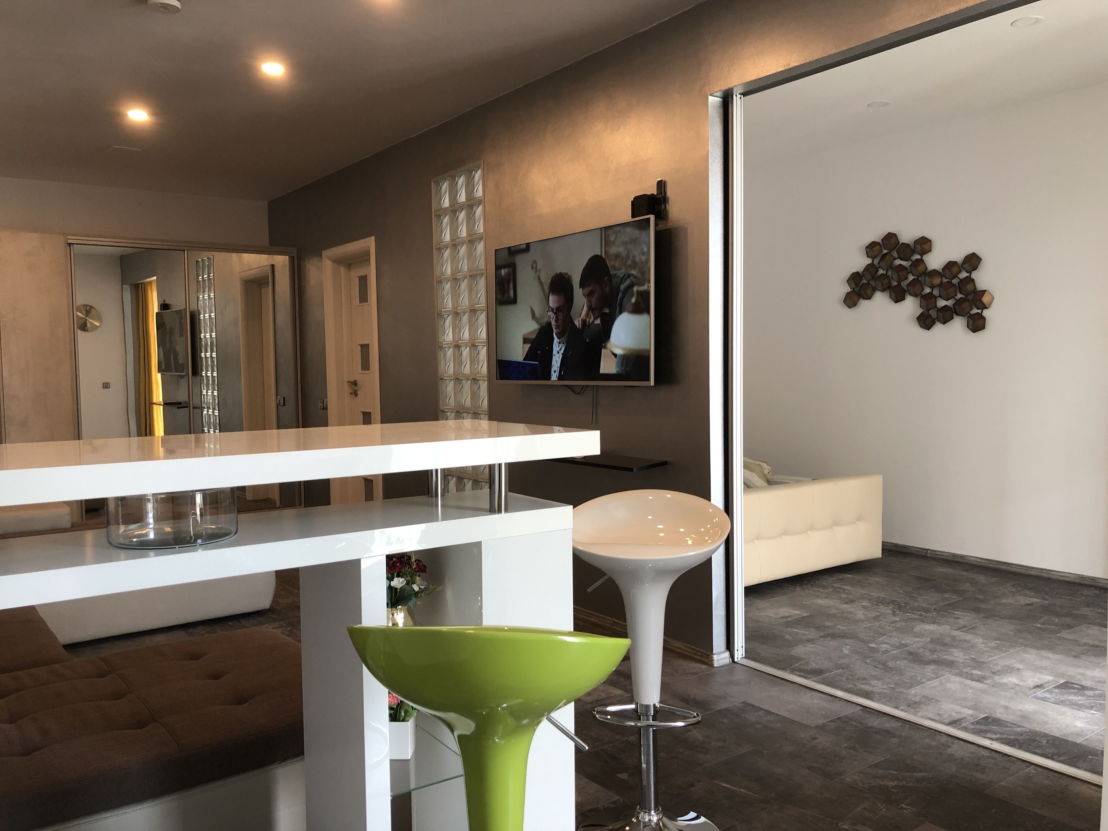 Un salon moderne avec un canapé blanc, un tabouret de bar vert, une table basse blanche et un téléviseur fixé au mur.