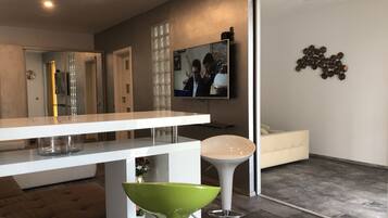 Un salon moderne avec un canapé blanc, un tabouret de bar vert, une table basse blanche et un téléviseur fixé au mur.