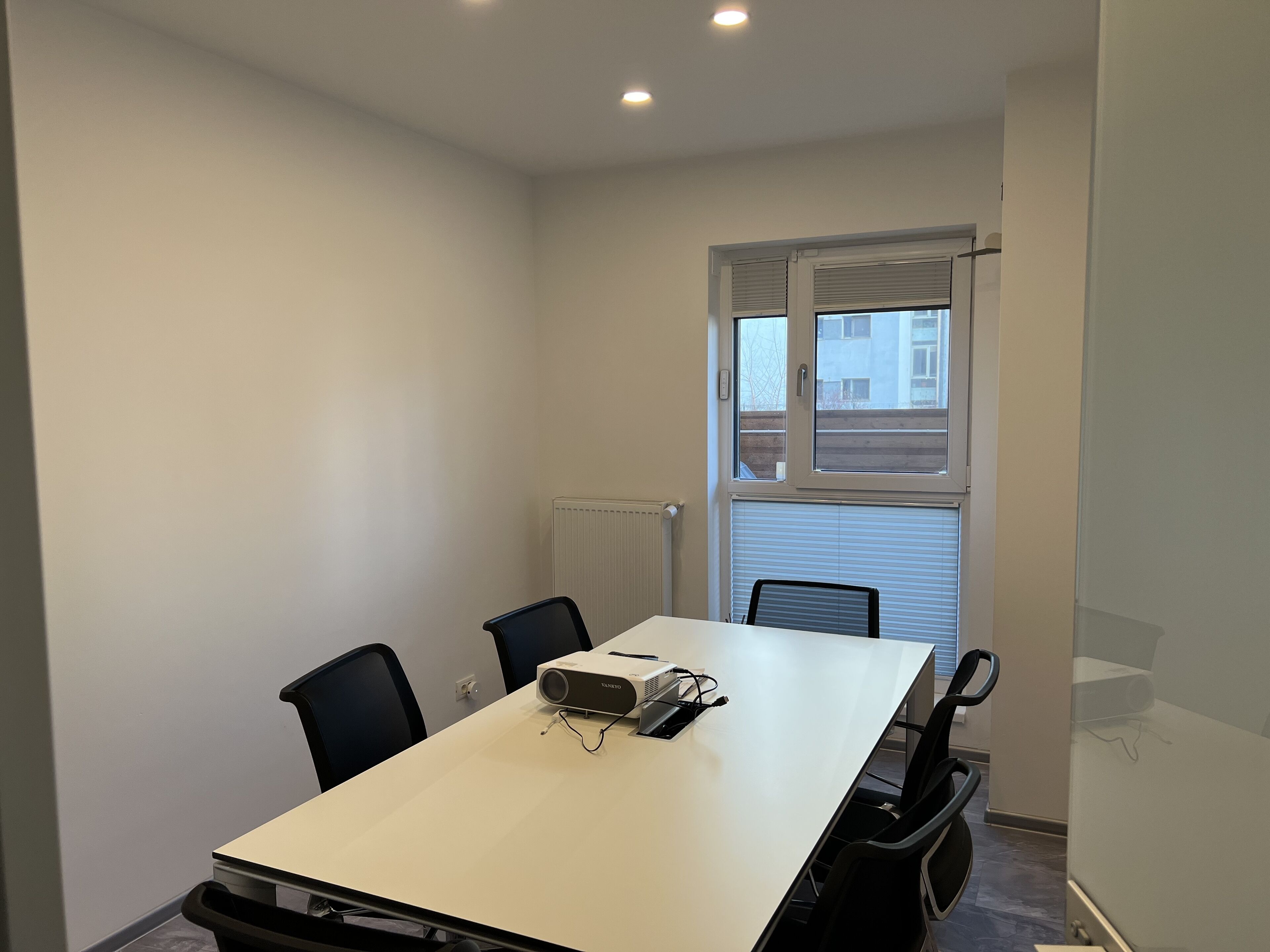 A conference room with a white table, four black chairs, a projector, and a window with blinds.