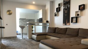 A modern living room with a sectional sofa, a kitchen area with white cabinets, and a marble backsplash. - Westpark 2 Residence Lakeview W6 (Bucharest)
