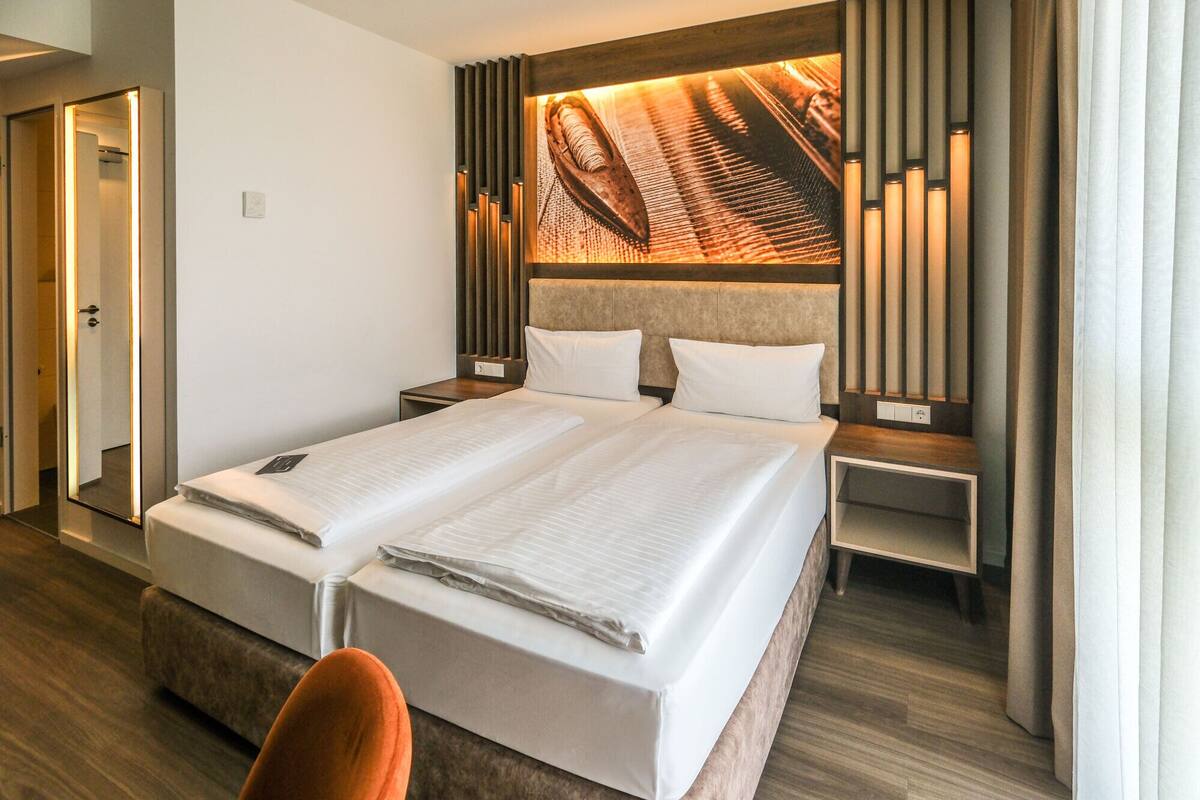 Un lit double avec une tête de lit ornée de lattes en bois verticales et dotée d’un panneau lumineux intégré. Il y a deux tables de chevet, l’une avec une lampe et l’autre avec une prise de courant. La pièce est dotée d’un plancher en bois et d’une porte qui donne sur un autre espace.