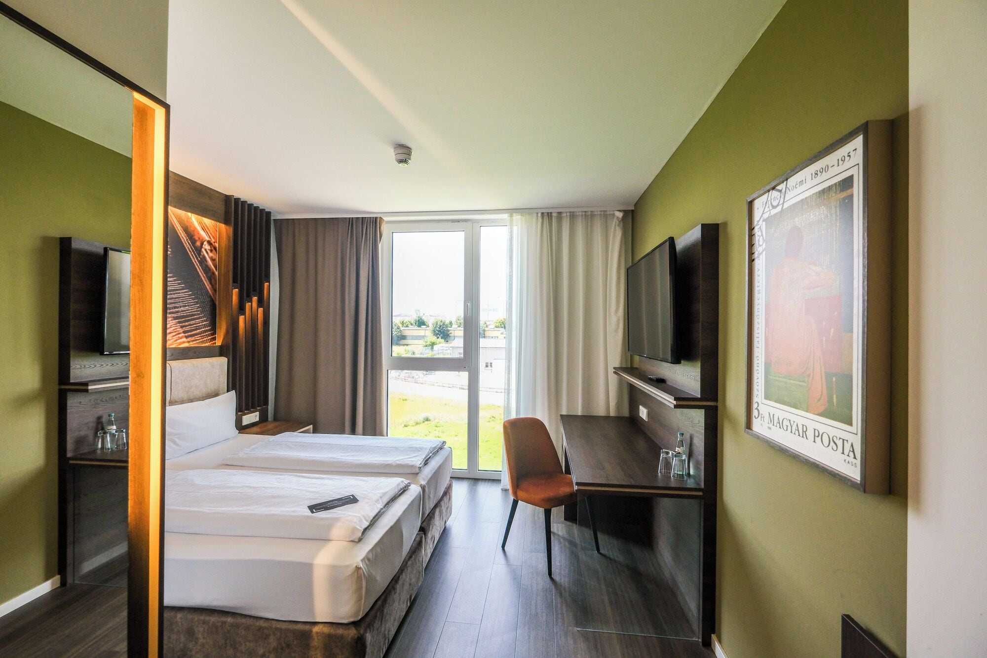 A hotel room with two beds, a desk, a chair, a television, and a large window.