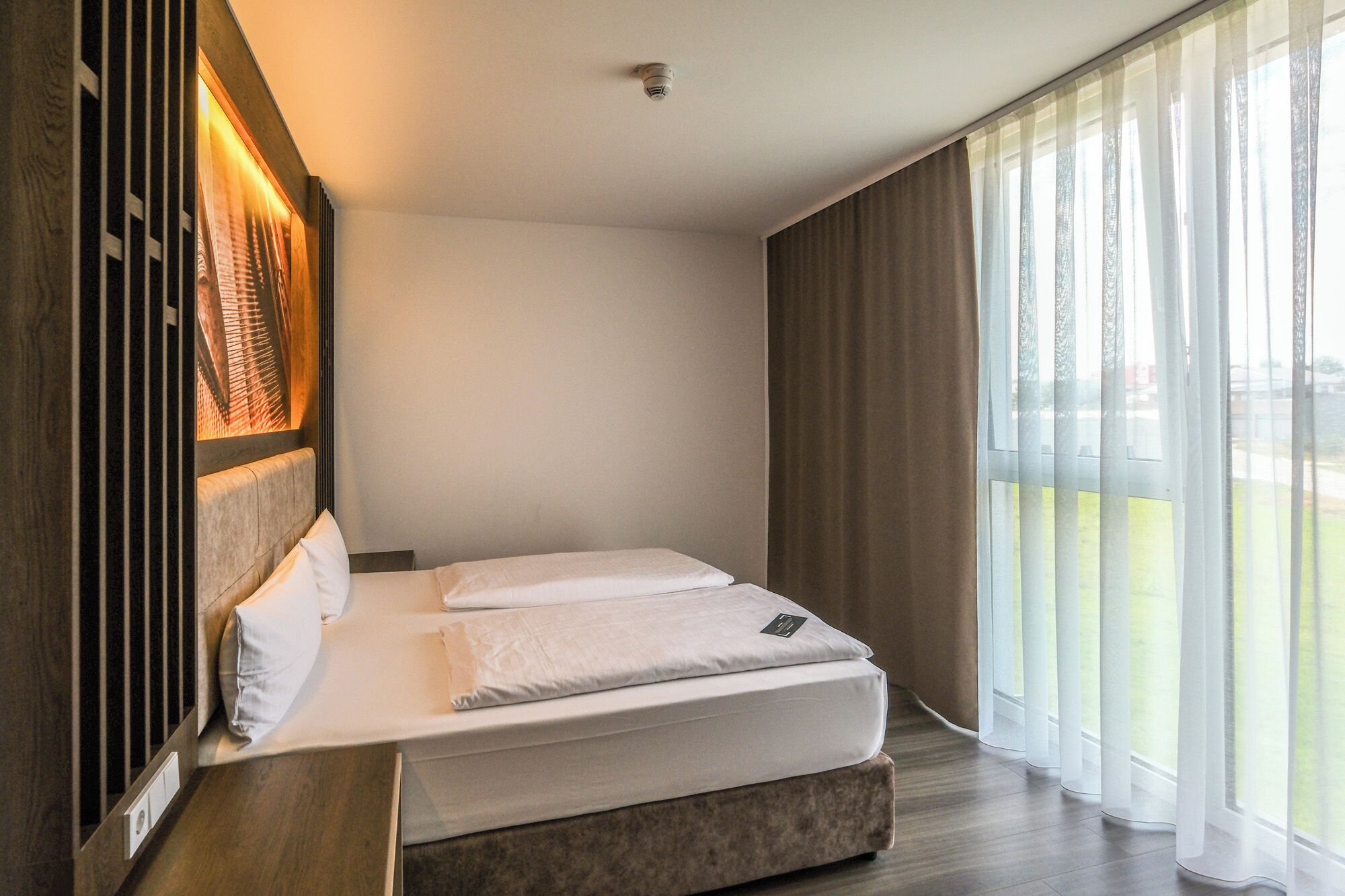A modern hotel room with a large bed, a view of the outdoors, and a wooden accent wall.