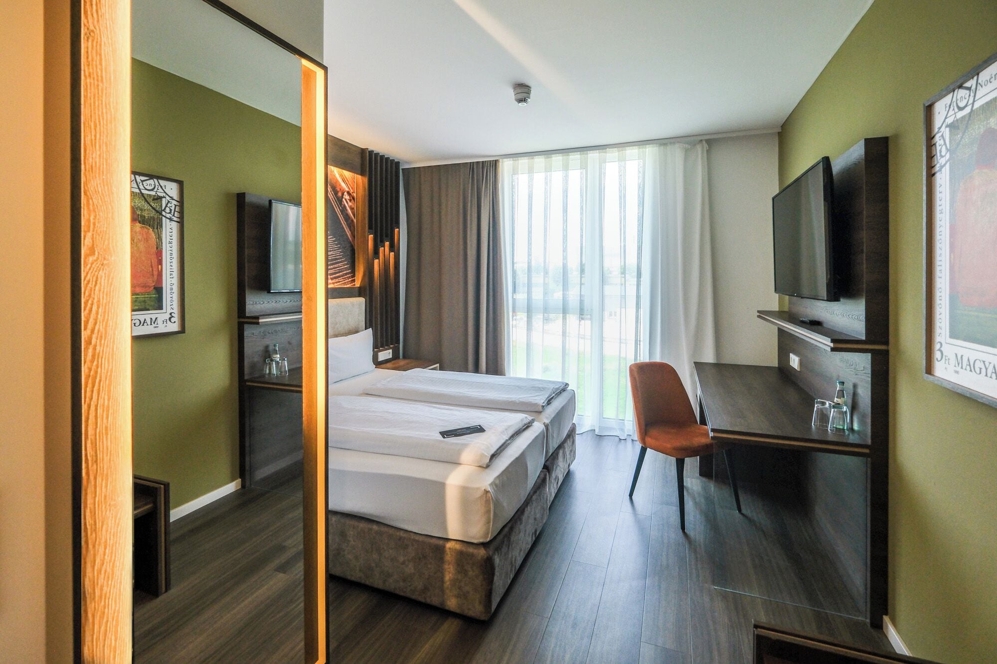 Ein Hotelzimmer mit Bett, Schreibtisch, Stuhl, Fernseher und einem großen Fenster mit Vorhängen.