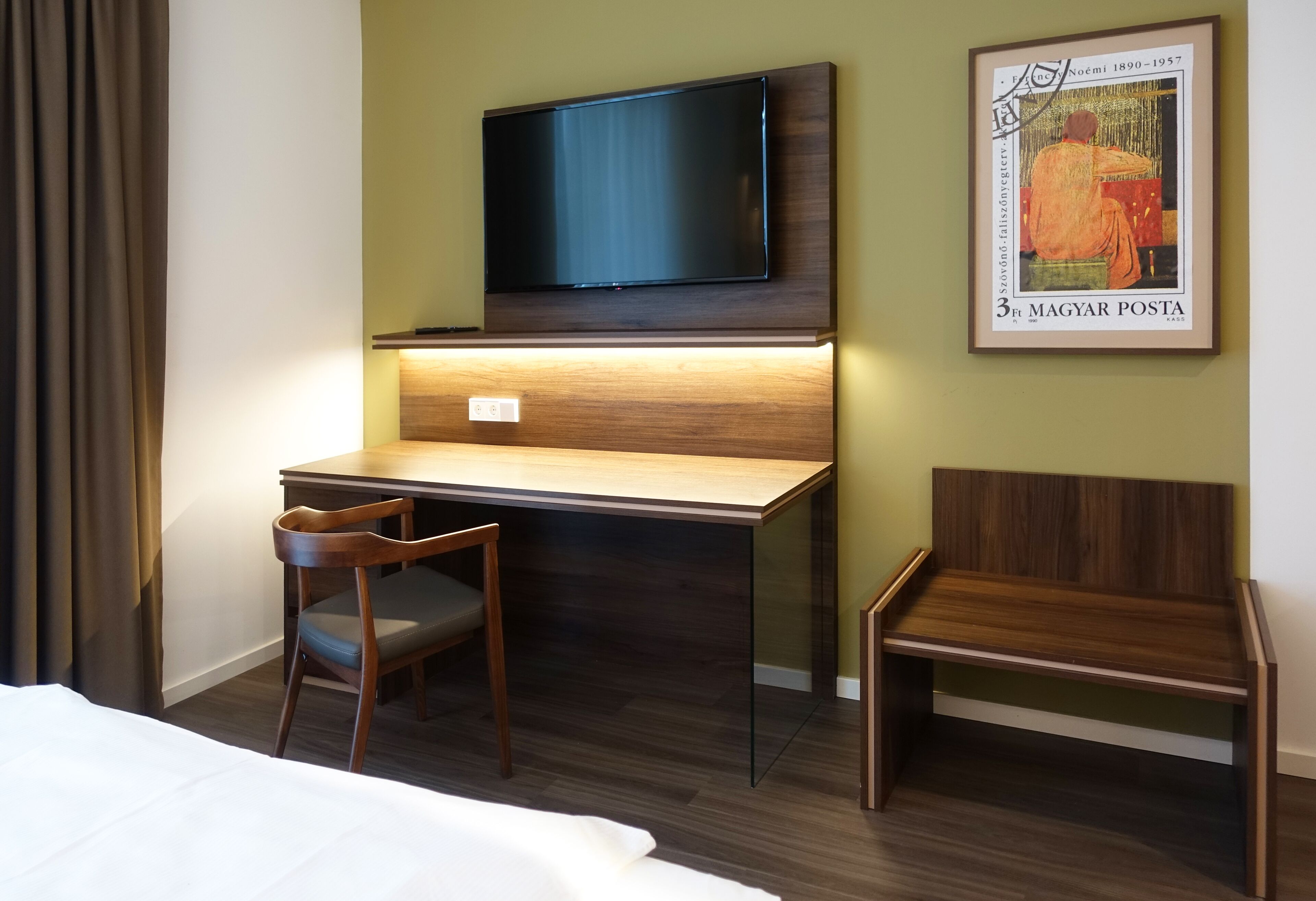 A hotel room with a wooden desk, a television mounted above it, a framed artwork on the wall, and a bed with white bedding.