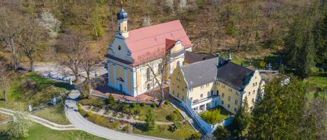 An aerial view of a large, historic building with a prominent tower and a red roof, surrounded by a well-maintained garden and a winding road.