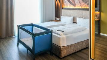 A hotel room with two beds, a blue playpen, wooden flooring, and a large window.