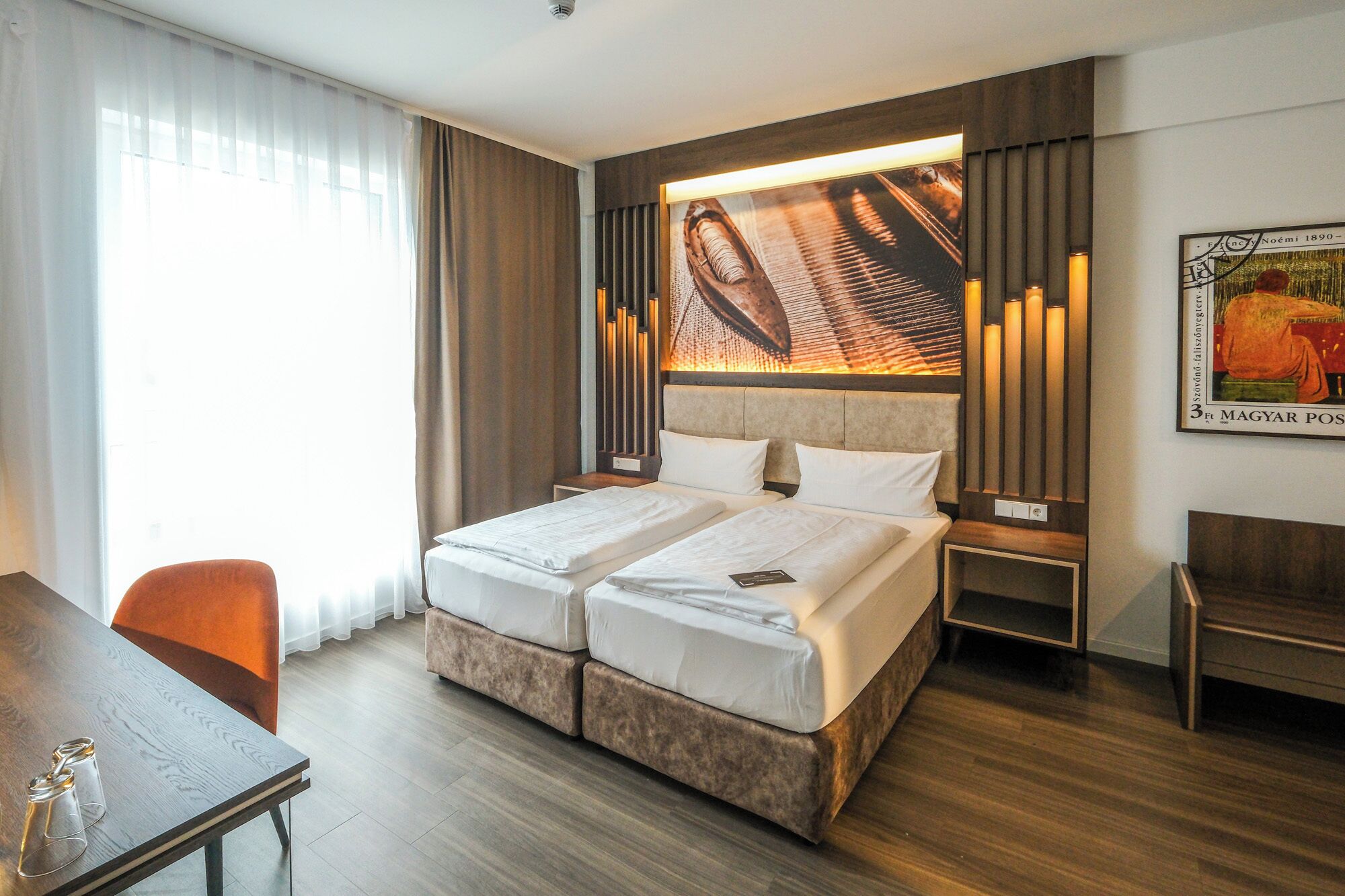 Ein Hotelzimmer mit einem Doppelbett, einem Schreibtisch mit zwei Stühlen, einem großen Fenster mit Vorhängen und einem Wandbild.