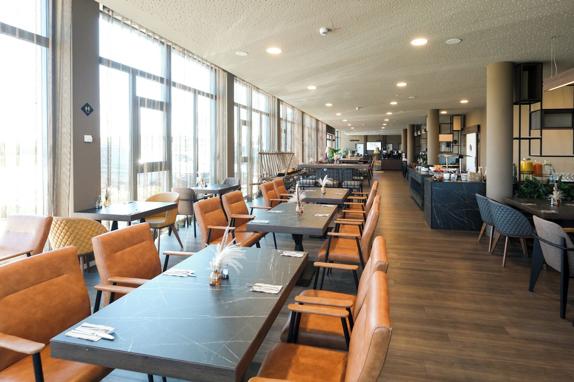 Un restaurant espaiós amb taules i cadires llargues, grans finestres i un disseny interior modern.