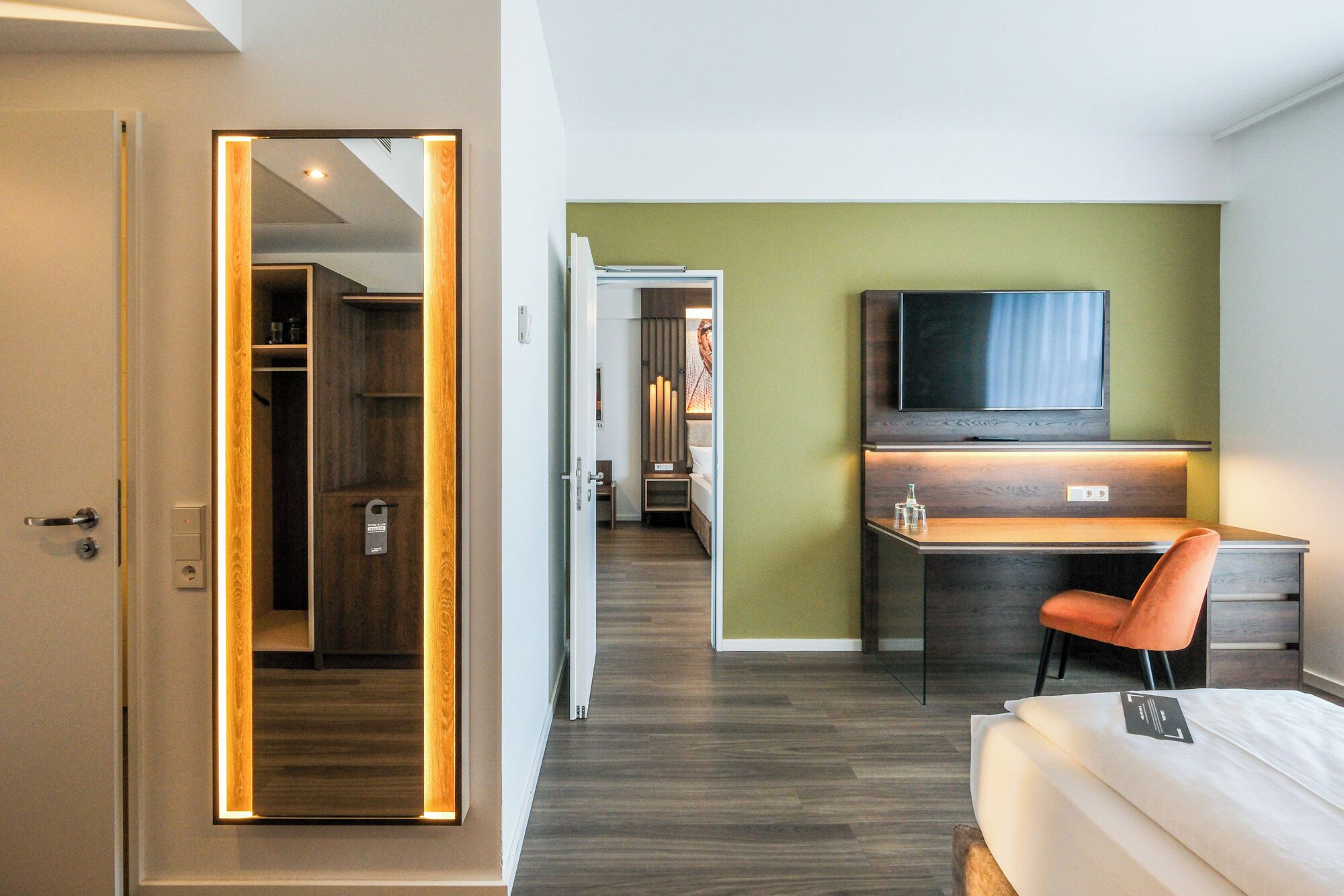 A hotel room with a wooden floor, a desk with a chair, a bed, a TV, and a wardrobe.
