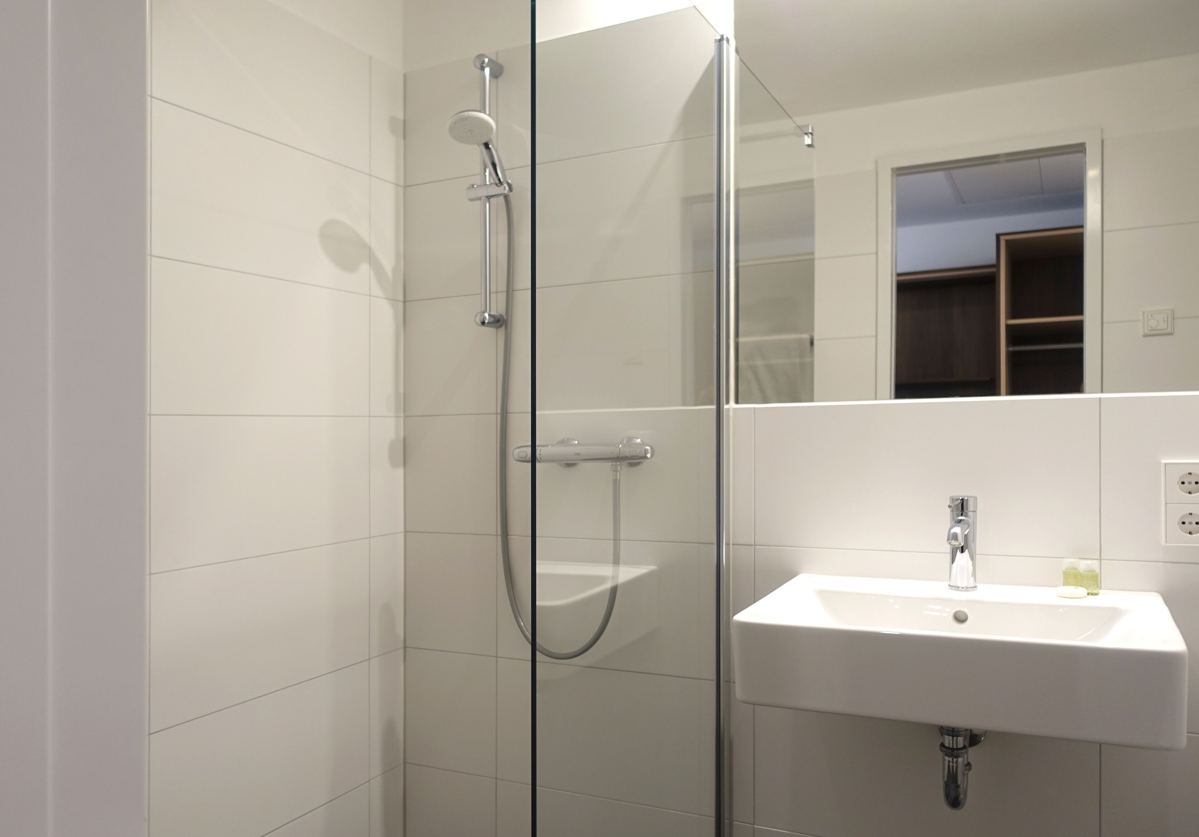 A modern bathroom with a glass-enclosed shower, a white sink, and a mirror reflecting a wardrobe.
