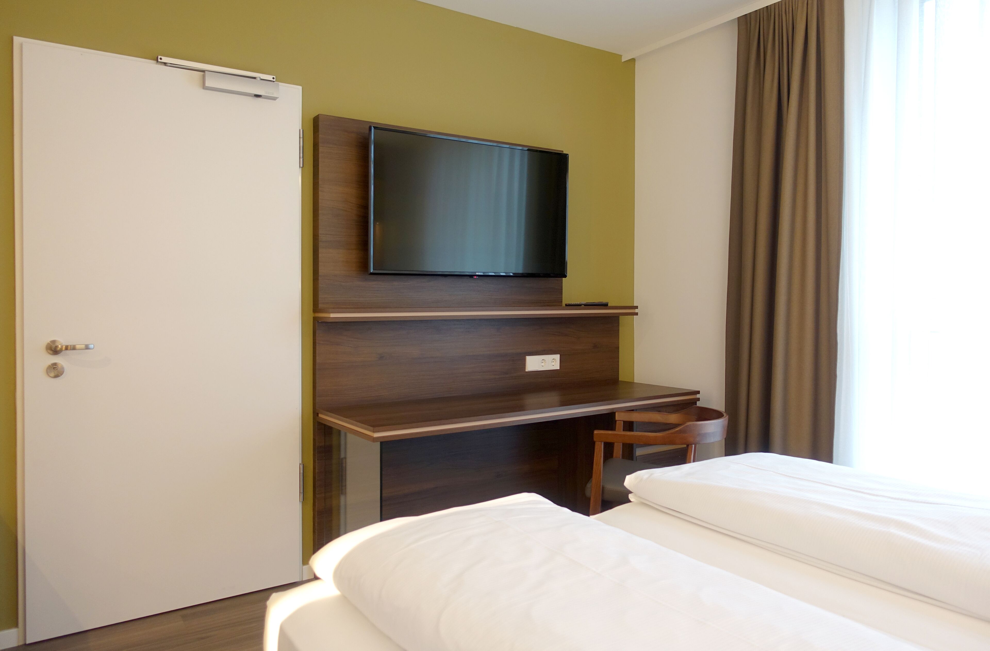 A hotel room with a bed, a desk, a television mounted on the wall, and a door.