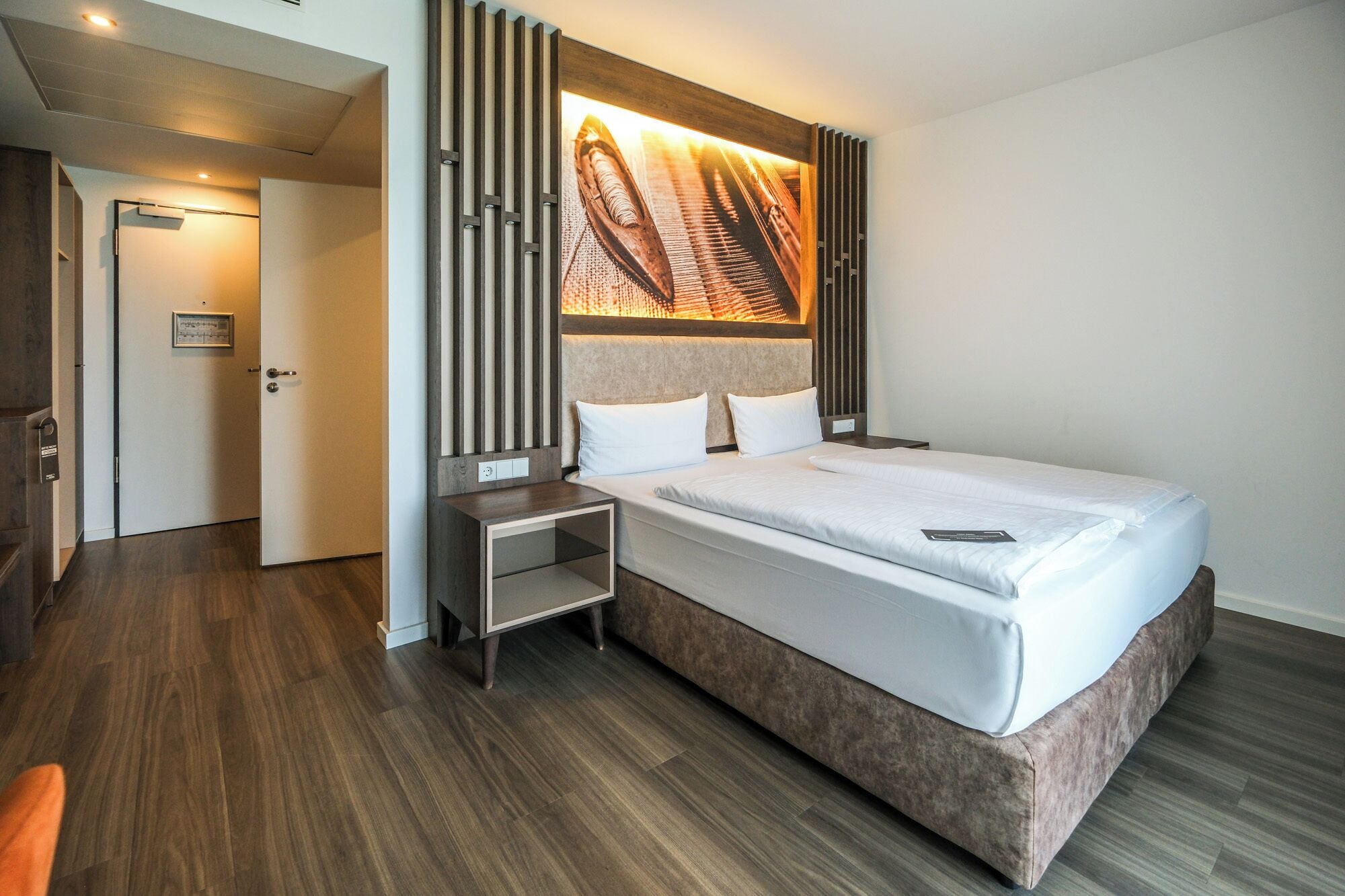 A modern hotel room with a large bed, a bedside table, and a headboard with a vertical light panel.