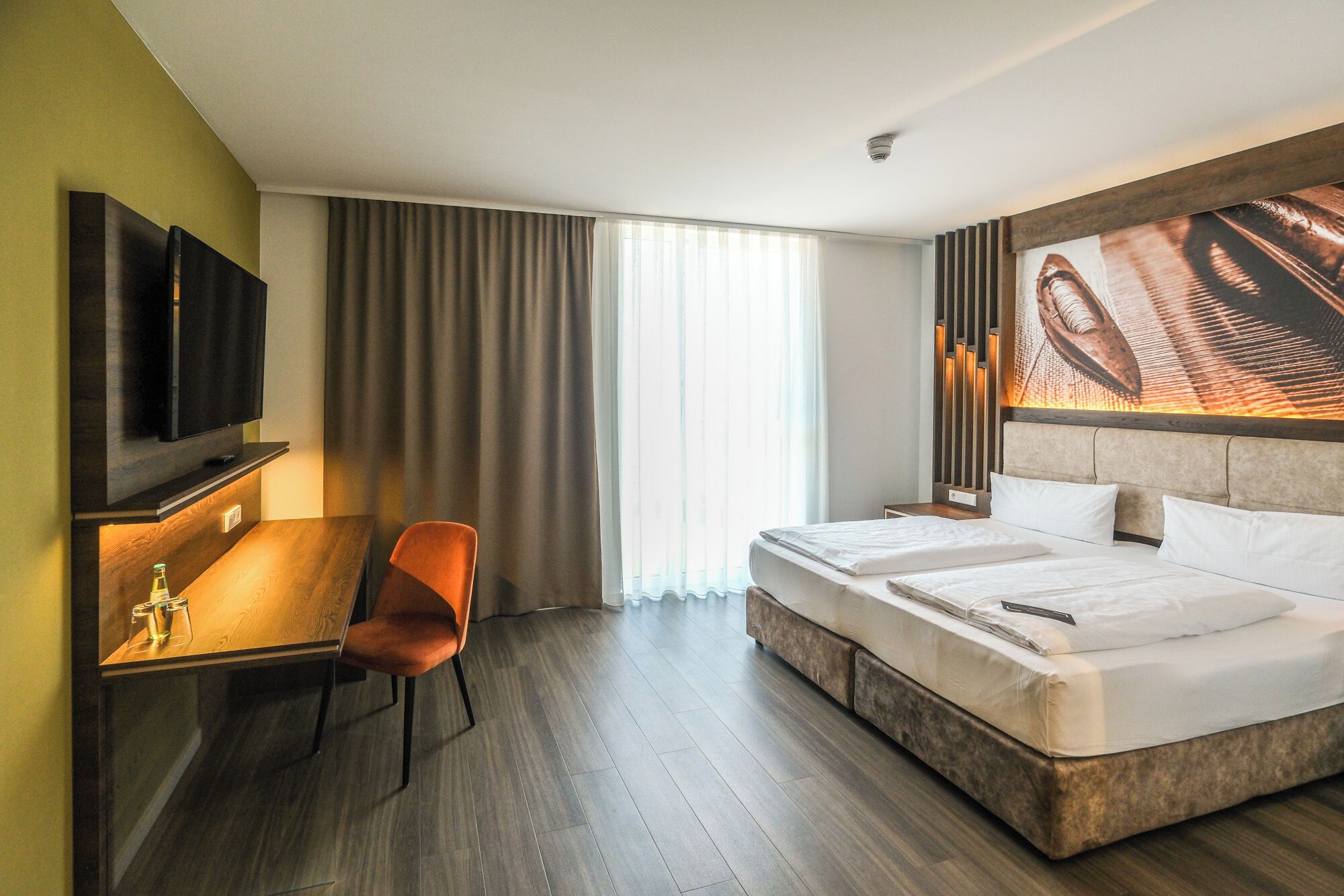 Hotelkamer met een bed, bureau, stoel, televisie en een groot raam met gordijnen.