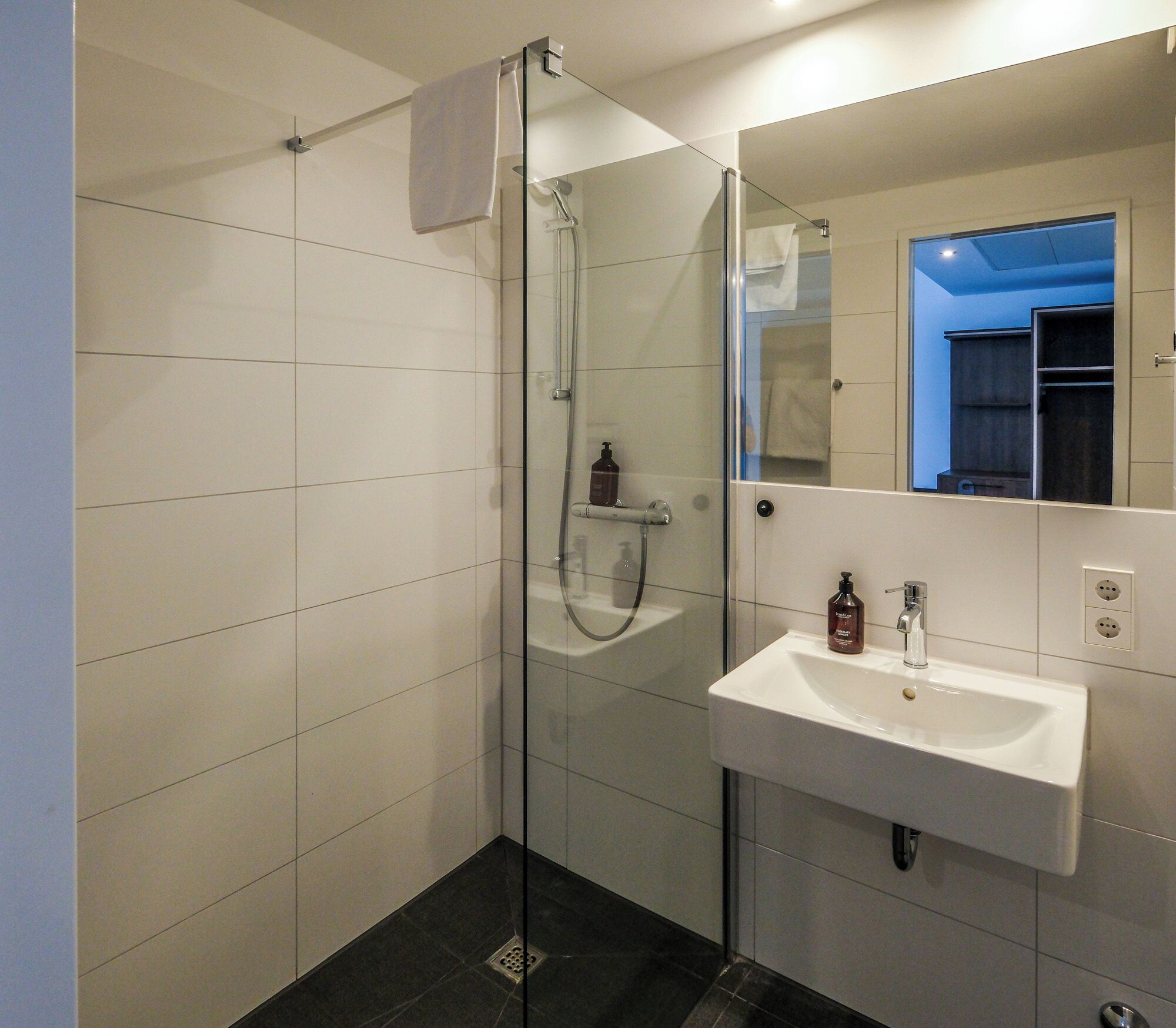 Un baño moderno con ducha acristalada, un lavabo blanco y un espejo que refleja una pared azul y un armario.