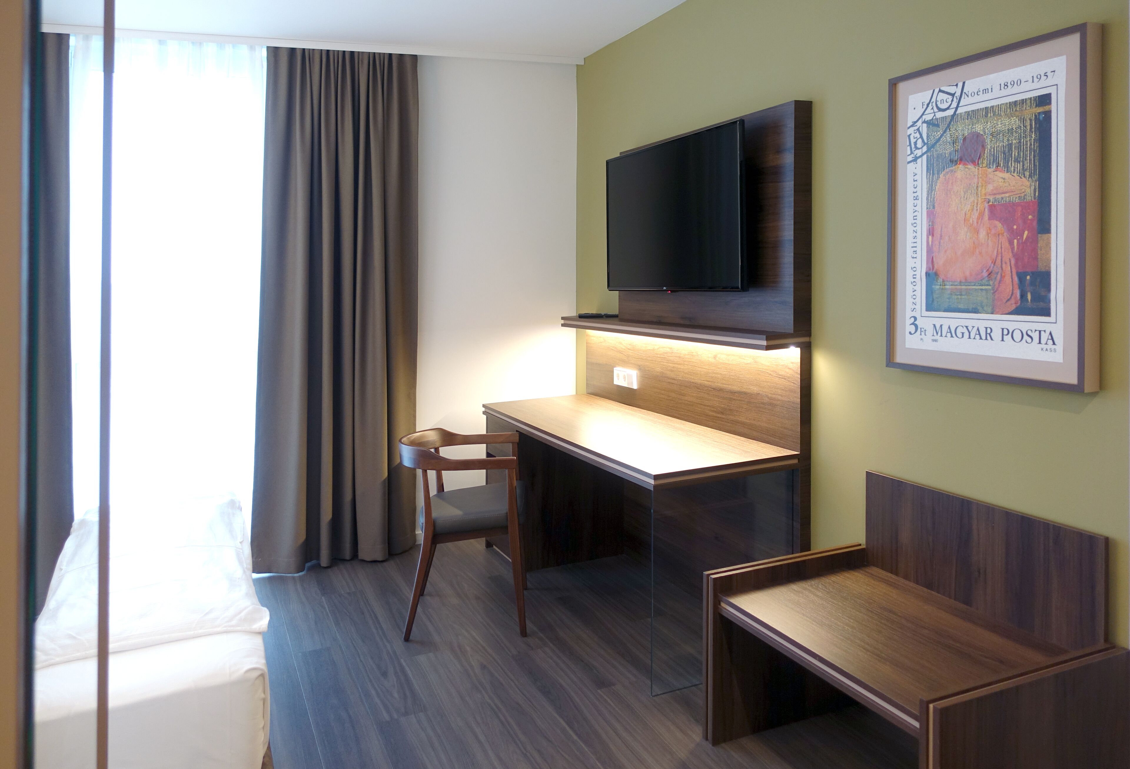 A hotel room with a bed, a desk with a chair, a television mounted on the wall, and a framed picture.