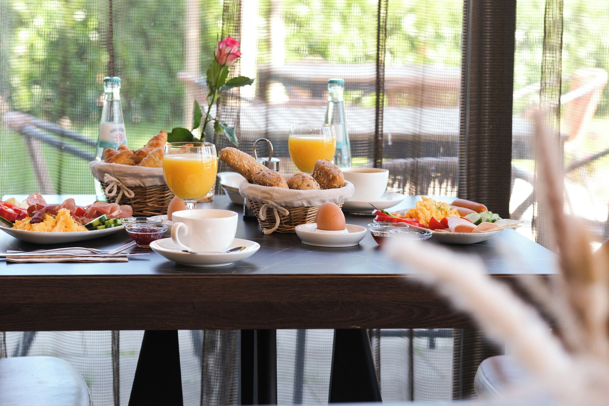 Een ontbijttafel met diverse etenswaren, waaronder brood, eieren en fruit, op een balkon met uitzicht op groen.