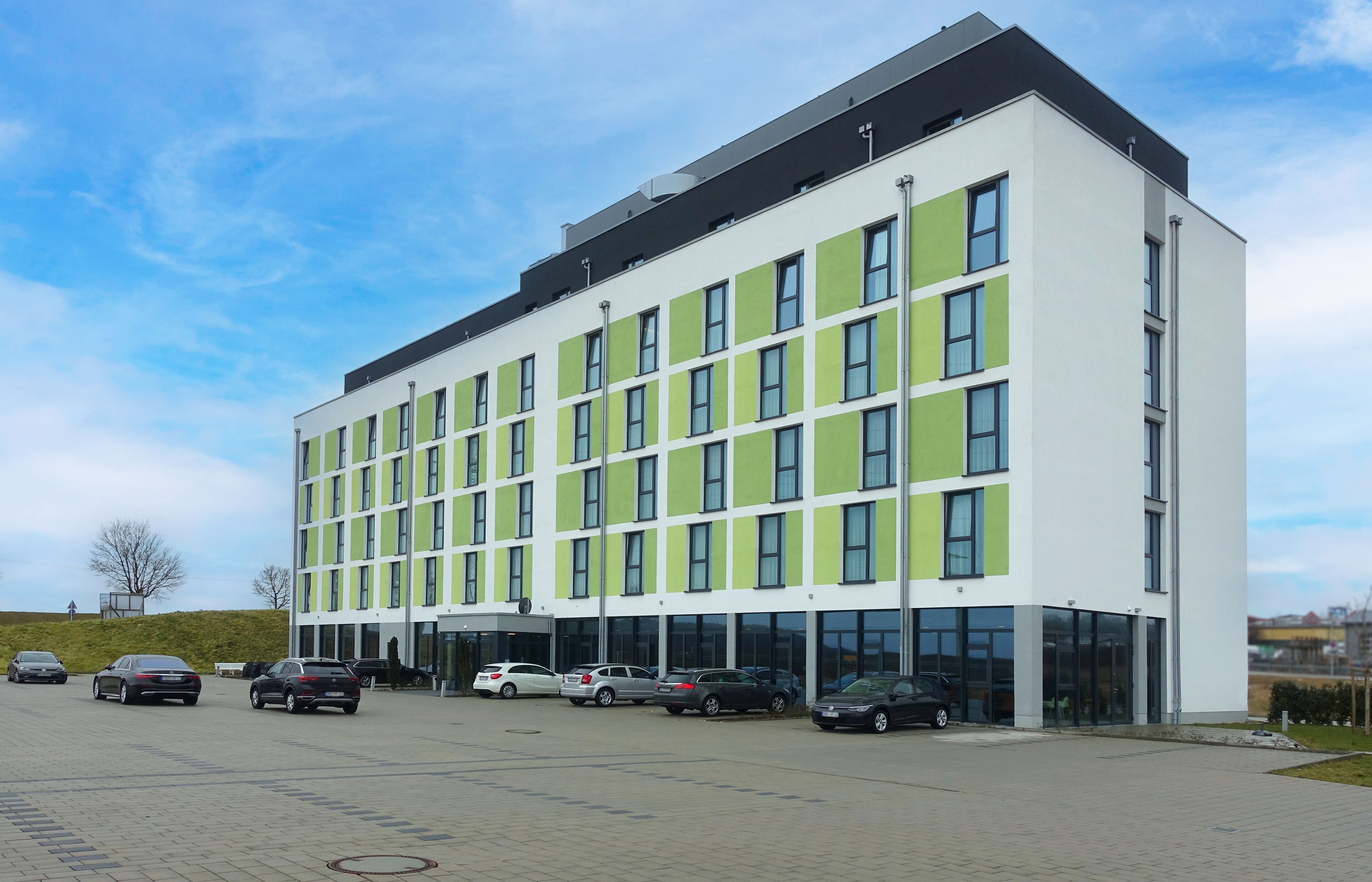 Un bâtiment moderne doté d’un toit plat, de grandes fenêtres et d’un parking où sont stationnées plusieurs voitures.