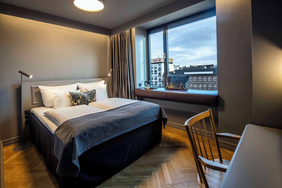 A hotel room with a bed, a desk, a chair, a window with a view of buildings, and a lamp.
