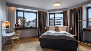 Una habitación de hotel moderna con una cama grande, un escritorio y una silla. Amplias ventanas con cortinas ofrecen vistas de edificios cercanos.
