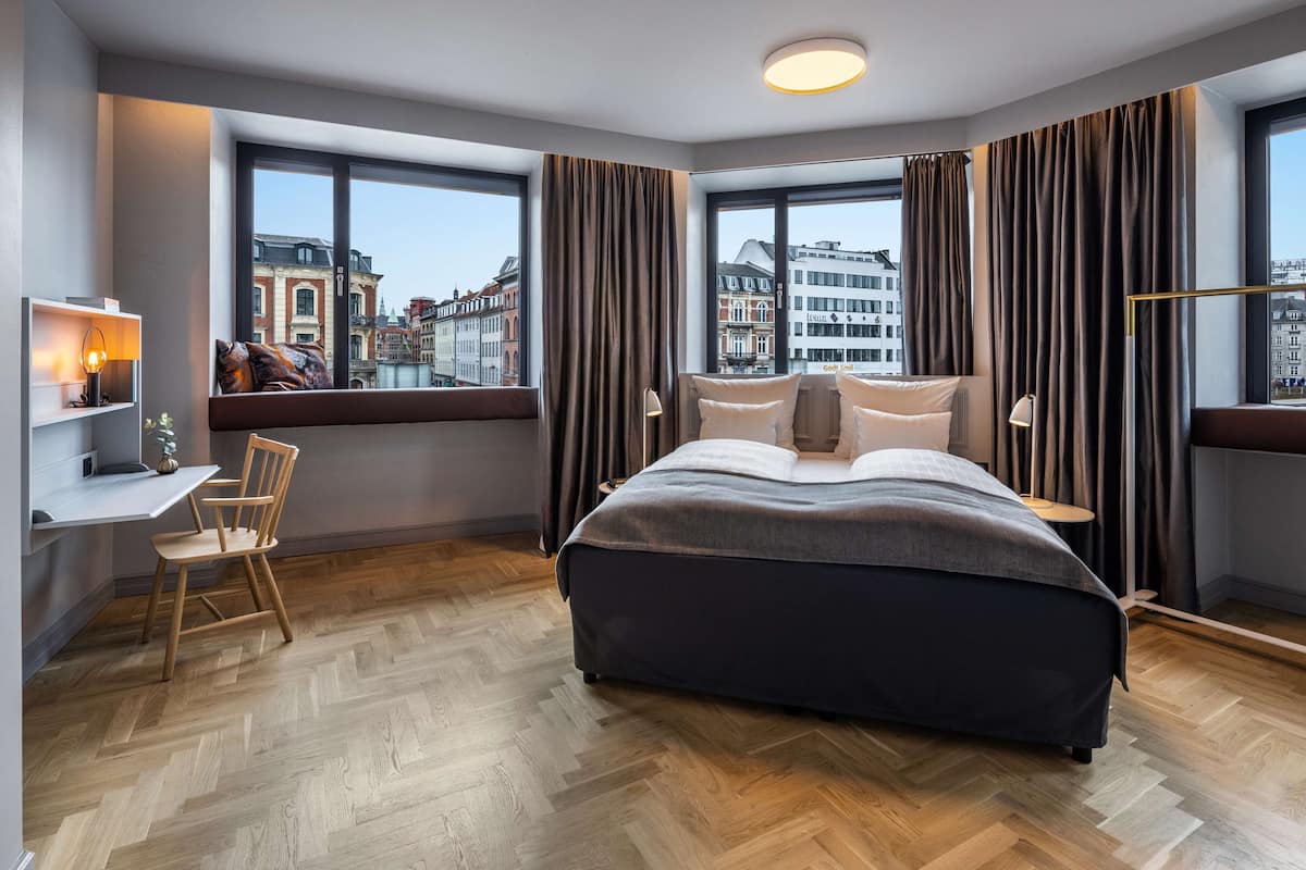 A modern hotel room with a large bed, a desk, and a chair. There are large windows with curtains and a view of buildings outside.