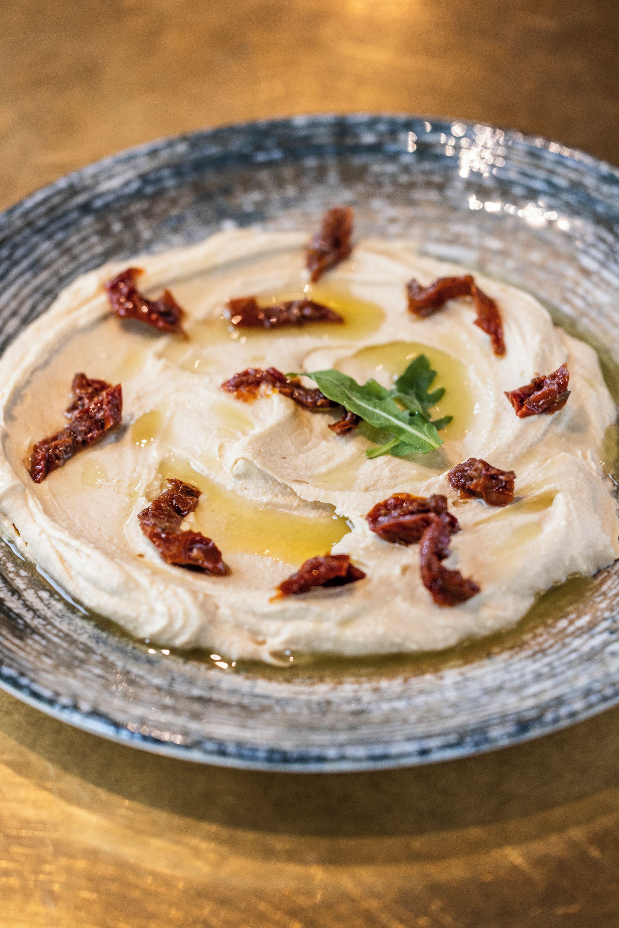 A bowl of creamy dip with olive oil and crispy bits on a textured plate.