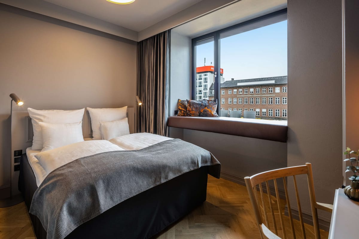 A modern hotel room with a large bed, a wooden chair, a small table, and a window with a city view.