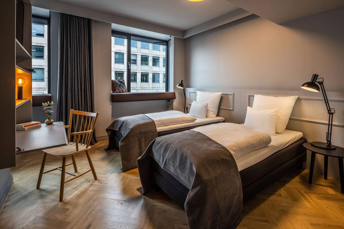 A hotel room with two beds, a desk, a chair, and a window with curtains.