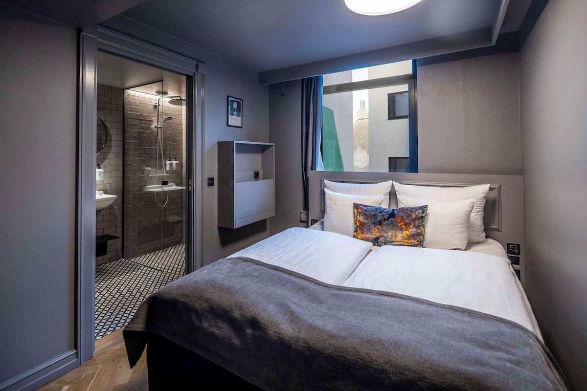A modern hotel room with a large bed, a bedside table, a mirror, and a view of the outside.