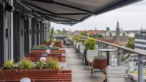 Una terraza al aire libre con bancos y sillas de madera, plantas en macetas y vistas a un paisaje urbano con edificios y una bandera.
