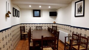 Un espace repas avec une grande table en bois entourée de chaises, un sol carrelé, un téléviseur fixé au mur et un tableau encadré.