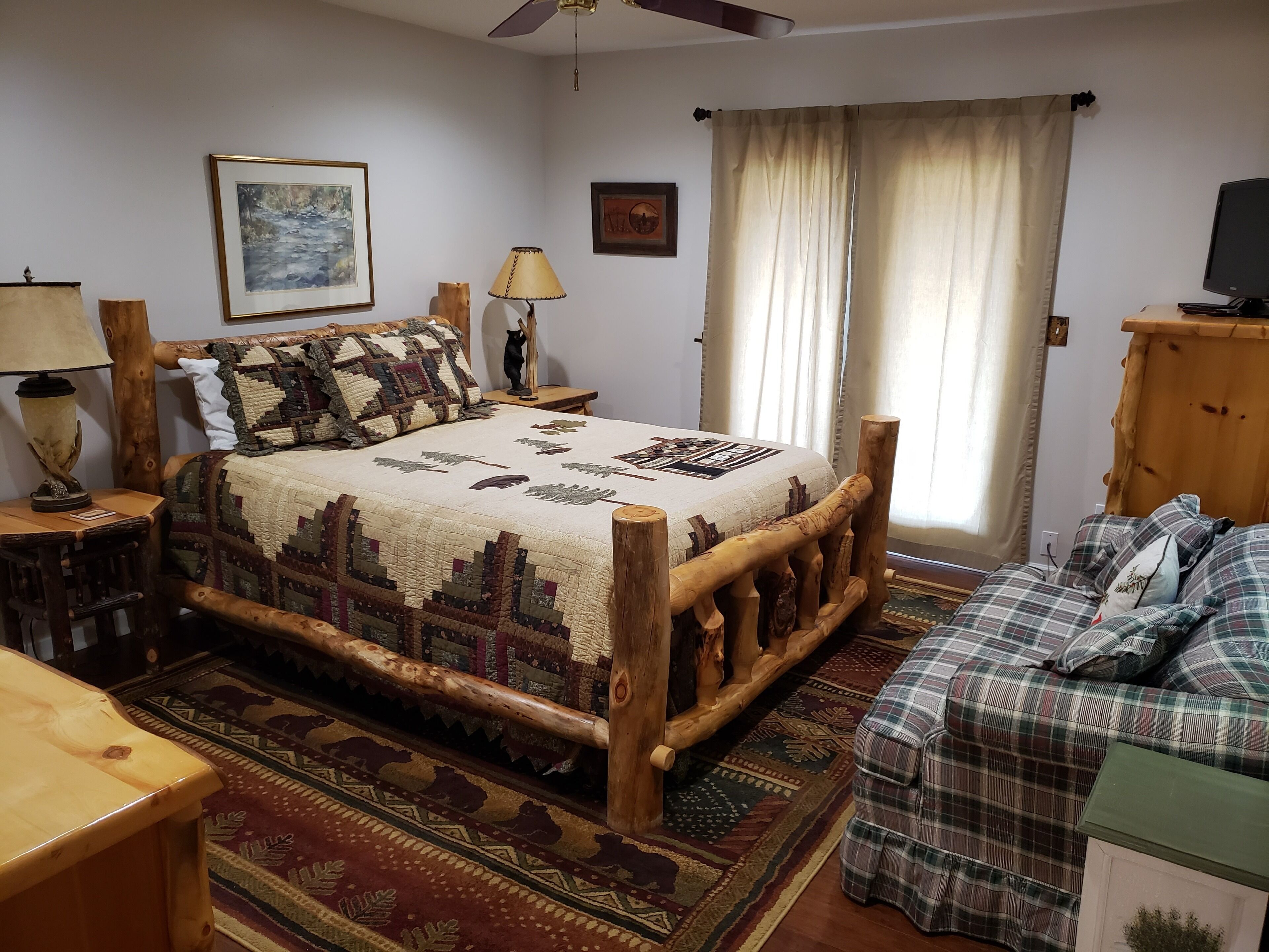 A bedroom with a wooden bed, a wooden nightstand, a lamp, a cat statue, a framed picture, a window with curtains, and a television on a wooden stand.