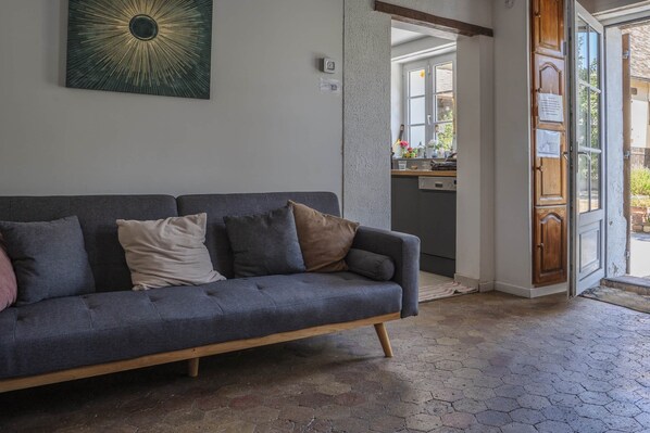 Un sofá gris con varios cojines, un área de cocina con lavavajillas y una puerta de madera que da al exterior.