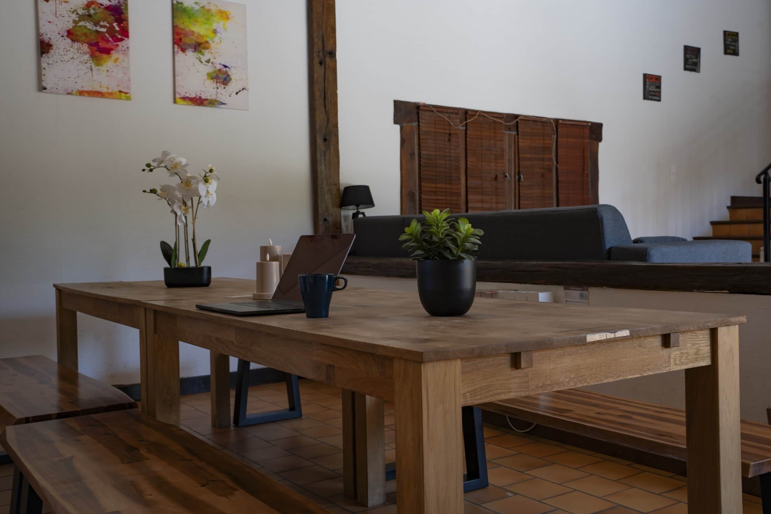 Una mesa de madera con una computadora portátil, una planta en maceta y un florero con flores en una habitación con piso de baldosas y un sofá al fondo.