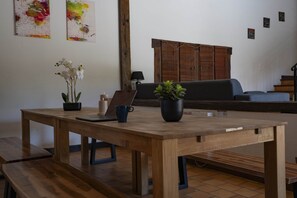 Une table en bois avec un ordinateur portable, une plante en pot et un vase de fleurs, dans une pièce dont le sol est carrelé et où l’on aperçoit un canapé au fond.