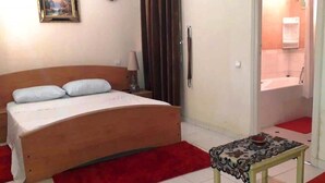 A bedroom with a wooden bed, a red rug, a small table with a decorative cover, a nightstand, a lamp, and a painting on the wall. - Residence Atlas Hivernage 4841 (Marrakech)