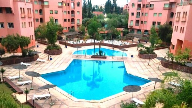 A swimming pool surrounded by lounge chairs and umbrellas, with a backdrop of pink buildings and greenery. - Residence Atlas Hivernage 4842 (Marrakech)