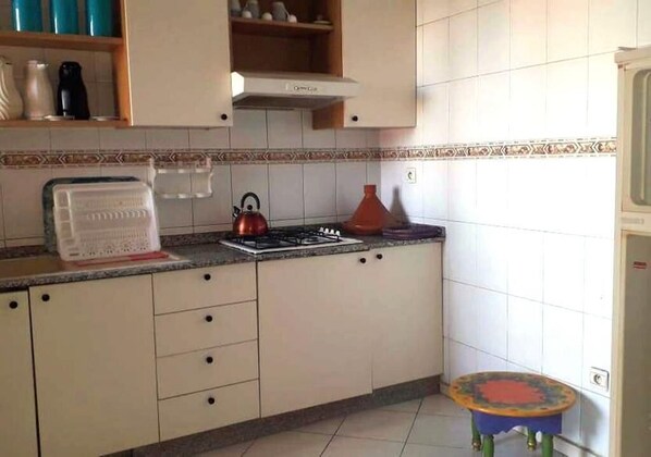 Une cuisine avec des meubles blancs, une cuisinière à gaz, une bouilloire et un petit tabouret.