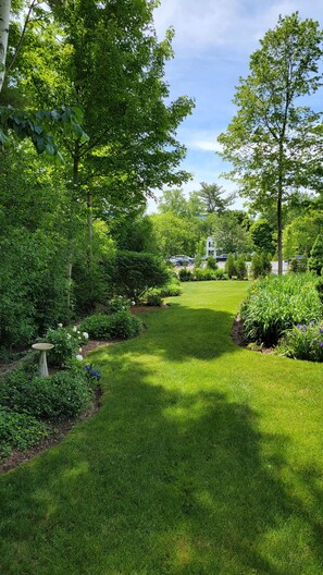A well-maintained garden with a variety of plants and a lawn, leading to a white house with a visible driveway.