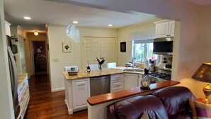 A modern kitchen with white cabinets, a granite countertop, and stainless steel appliances. There is a dining area with a leather sofa and a lamp.