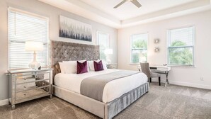 Une chambre avec un grand lit, un bureau et une chaise. Il y a deux fenêtres équipées de stores, un ventilateur de plafond et une horloge fixée au mur.