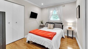 A bedroom with a bed, a TV, a window with curtains, a bedside table with a lamp, and a wardrobe. - Hot Tub | Sauna | Fire Pit | Outdoor Living | Elegant Ranch (Ann Arbor)