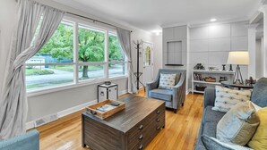 A modern living room with a wooden coffee table, a grey sofa, and a blue armchair. There is a large window with curtains, a bookshelf, and a lamp on a side table. - Hot Tub | Sauna | Fire Pit | Outdoor Living | Elegant Ranch (Ann Arbor)